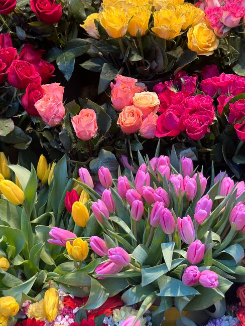 A vibrant floral display featuring a variety of fresh flowers arranged closely together. Prominently, there are pink and yellow tulips with elongated green leaves, complemented by clusters of pink, pe